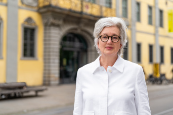 Eine Frau mit weißgrauen kurzen Haaren, einer runden dunklen Brille und weißer Bluse vor einem Gebäude