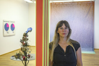 Eine Frau mit Brille und langen blonden Haaren steht in einem Raum. Auf einem Tisch steht eine Vase. 