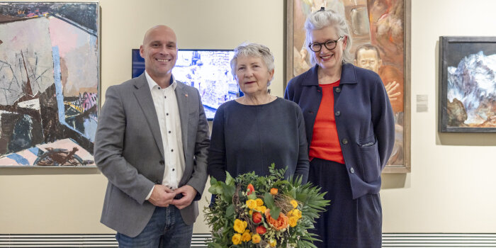 zwischen einem Mann und einer Frau steht eine Frau mit kurzen grauen Haaren und BLumenstrauß