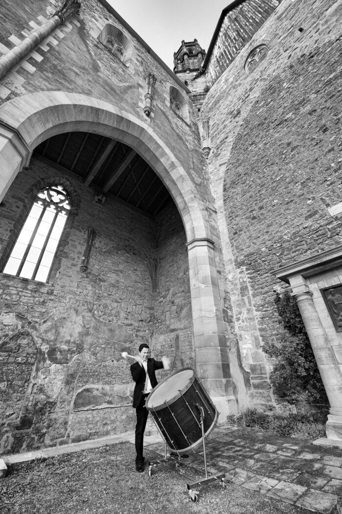Schwarz Weiß Aufnahme eines alten Gebäudes, im Vordergrund ein Mann im Anzug und eine Trommel