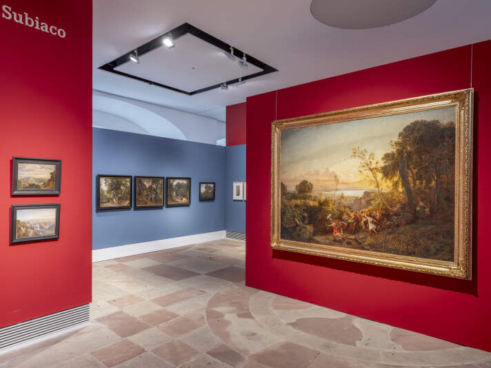 Ein Ausstellungsraum mit roten und blauen Wänden an den Gemälde hängen.