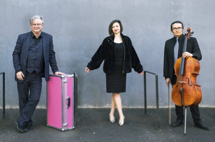 Drei Personen vor einer grauen Wand stehend mit einem Instrument und einem pinken Koffer