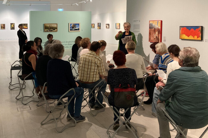 tea time with Barbara Eine Gruppe von Personen sitzt in einer Ausstellung, sie schauen auf eine Frau die etwas erklärt