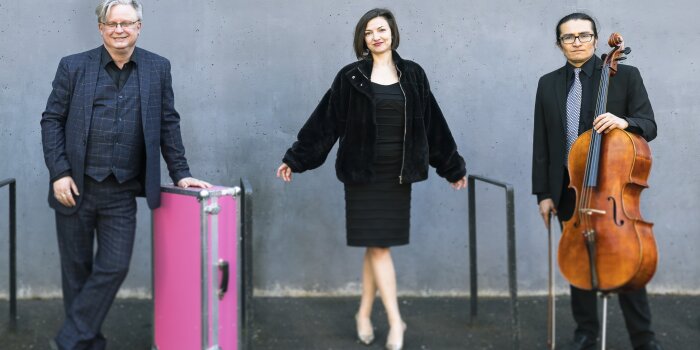 Drei Personen vor einer grauen Wand stehend mit einem Instrument und einem pinken Koffer