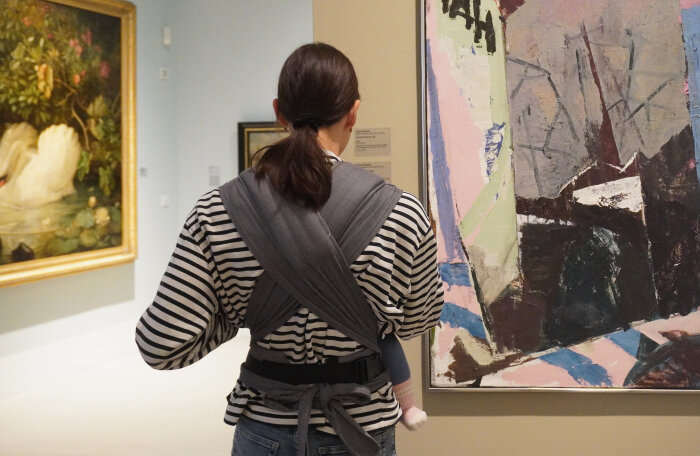 Eine Frau steht vor einem gemalten Bild, man sieht ihren Rücken und sie trägt ein Baby in einem Tragetuch