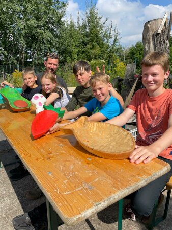 Kinder und ein Mann an einem Holztisch, die Kinder halten ihre Holz-Kunstwerke in ihren Händern