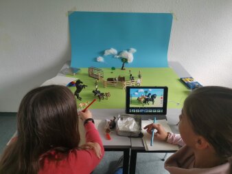 Kinder mit Stiften vor einer analogen Projektions-Fläche mit Koppel, Tieren und Wolkenhimmer-Hintergrund an der Wan