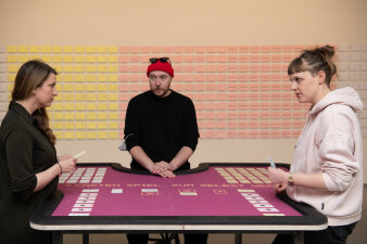 Zwei Frauen und ein Mann stehen an einem Spieltisch, im Hintergrund kleine gelbe und rosa Karten an einer Wand.