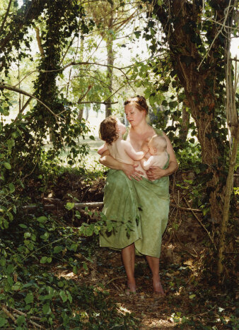 Foto einer jungen Frau mit nacktem Oberkörper und grünem Tuch mit zwei kleinen Kindern auf dem Arm in einem Wald