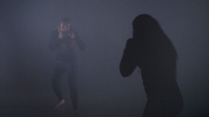 Person mit erhobenen Fäusten in einem dunklen Raum, rechts ein Schatten der Person
