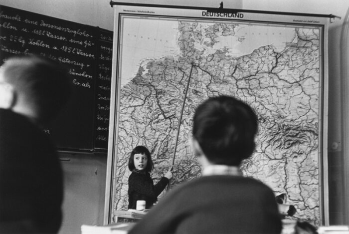 Titel: Schwarz weiß Foto das ein Kind vor einer Landkarte in einem Raum zeigt, im Vordergrund zwei unscharfe Personen 