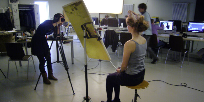 Ein Raum mit zwei Personen im Vordergrund, eine Person fotografiert eine andere sitzende Person