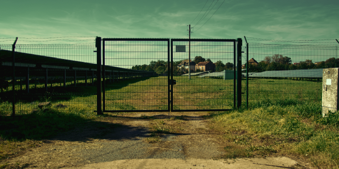 Ein Foto eines Weges mit Gras links und rechts, in der Mitte ein verschlosses Metalltor, im Hintergrund Gebäude