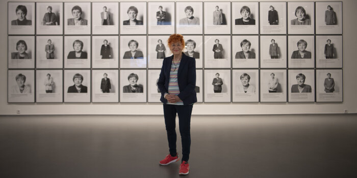 eine Frau mit roten Haaren steht vor einer Wand, an der Schwarz-Weiß-Fotografien von Angela Merkel hängen