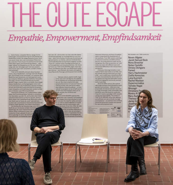 zwei Personen sitzen vor einer Wand mit Schriftzug "The Cute Escape" und langem Text