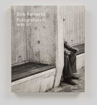 Foto: Katalog „Dirk Reinartz. Fotografieren, was ist“ © Steidl Verlag Schwarz-Weiß-Foto einer Person die hinter einer Mauer sitzt, vorne links weißer Text „Dirk Reinartz. Fotografieren,