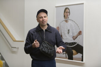 ein Mann mit dunkler Kleidung und Basecap steht gestikulierend vor einer gerahmten Fotografie