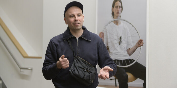 ein Mann mit dunkler Kleidung und Basecap steht gestikulierend vor einer gerahmten Fotografie