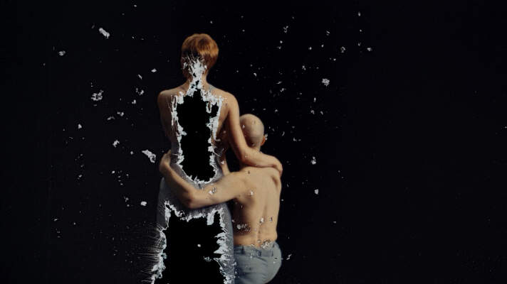 Eine stehende Person die sich aufzulösen scheint, daneben eine zweite Person sitzend