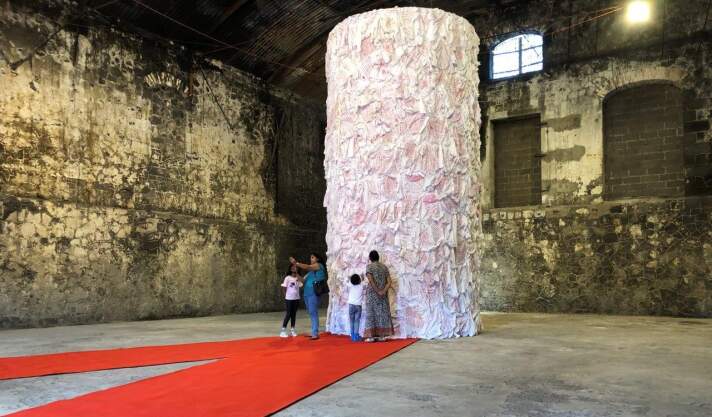 Column of Respect, 2019, Metallkonstruktion, beschriftete Blusen, roter Teppich, Aapravasi Ghat World Heritage Site & Cerne Docks, Port Louis (Mauritius) eine mit weißen Stoff behangene Säule in einem dunklen Raum, davor mehrere Personen stehend