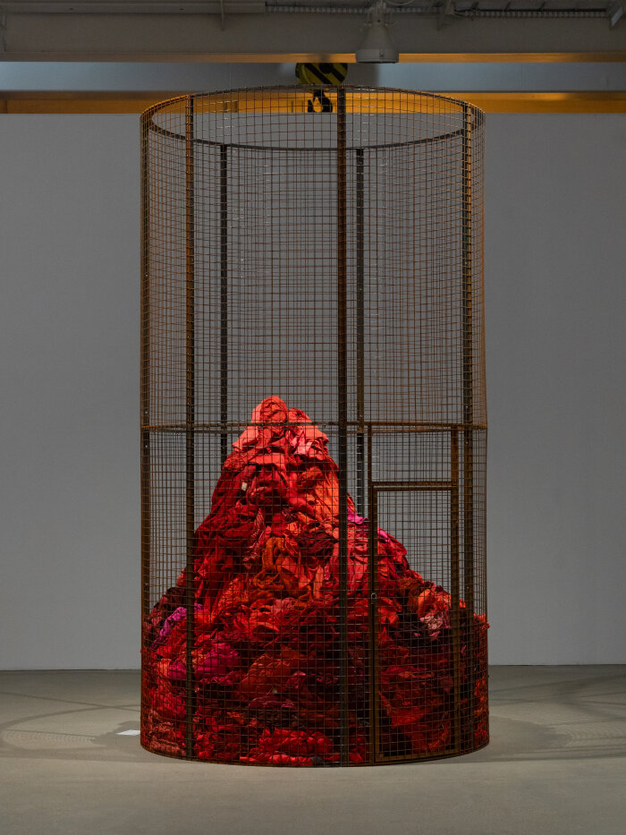 Eine runde rostige Metallkonstruktion in einem Raum stehend, darin rote gestapelte Stoffe