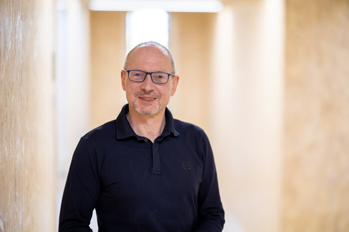 Ein lächelnder Mann mit Brille und dunklen Hemd vor einer hellen Wand