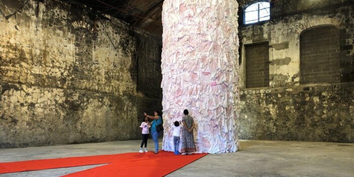 eine mit weißen Stoff behangene Säule in einem dunklen Raum, davor mehrere Personen stehend
