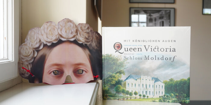 ein eingeschweißtes Buch steht neben einer Papiermaske, die eine Frau mit Blumenkranz abbildet