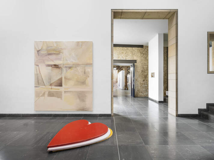 Blick in einen offenen Räum mit Gemälden, im Vordergrund ein herzförmiges Element auf dem Boden.