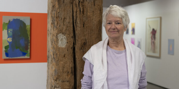 eine Frau mit kurzen Grauen Haaren steht in einer Ausstellung mit Grafiken an weißen Wänden und großem Holzbalken