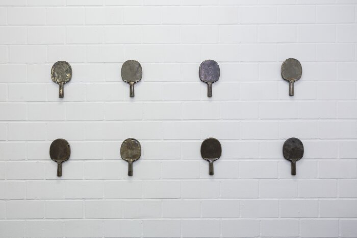 Tischtennisschläger aus einem dunklen Material in zwei Reihen an einer weißen Wand hängend