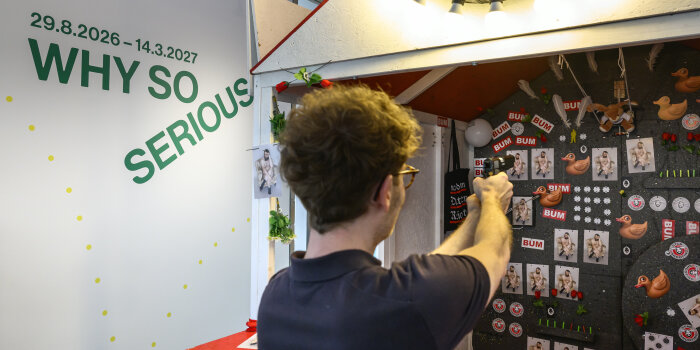 Ein Mann steht mit erhobener Waffe vor einer Schießbude, im Hintergrund an der Wand der Schriftzug „Why so serious“